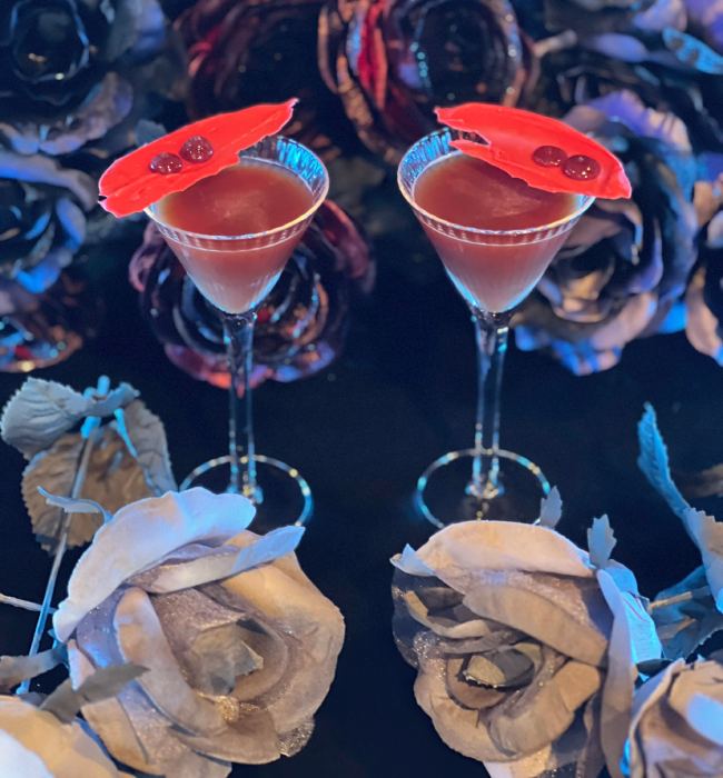 Two elegant red cocktails garnished with red petals, surrounded by dark and silver roses.