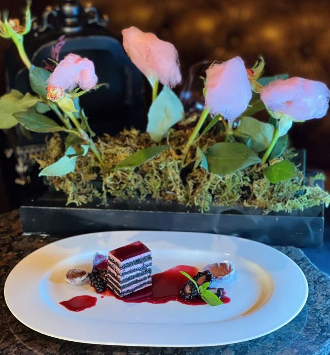 Plated layered dessert with berry sauce and blackberries, set in front of pink cotton candy roses and green foliage decoration.