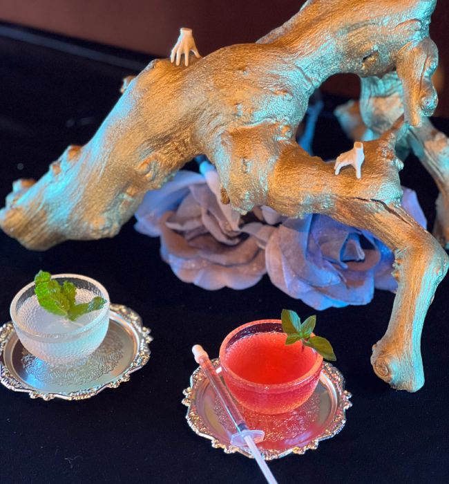 Two colorful cocktails on silver trays with mint garnish, placed beside a golden driftwood centerpiece and decorative flowers.
